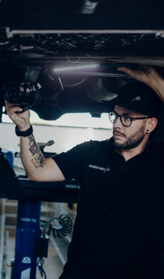 Techniker mit Brille und Tattoo, der eine Kamera unter einem Fahrzeug hält.