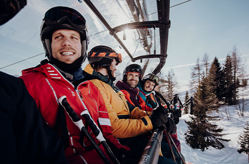 Vier Skifahrer sitzen in einem Sessellift und lächeln in die Kamera.