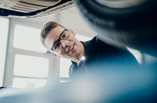 Junger Mechaniker mit Brille, der unter ein Fahrzeug schaut und eine Taschenlampe hält.