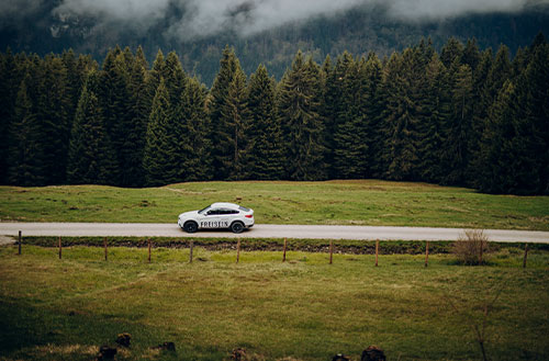 Ein weißes Auto fährt auf einer Landstraße entlang einer bewaldeten Landschaft.