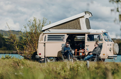 Ein beige Volkswagen Campingbus mit Aufstelldach steht in einer Landschaft, zwei Personen sitzen davor.