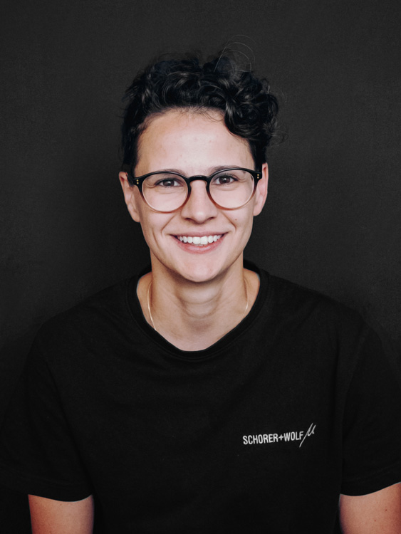 Junge Person mit Brille und lockigem Haar, die in die Kamera lächelt.