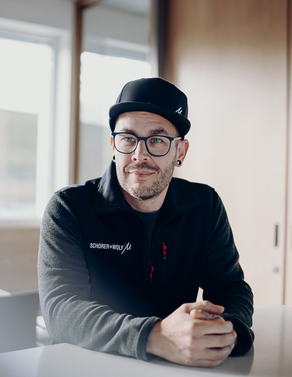 Mann mit Brille und schwarzer Mütze sitzt an einem Tisch, trägt einen grauen Pullover mit Logo von SCHORER + WOLF.