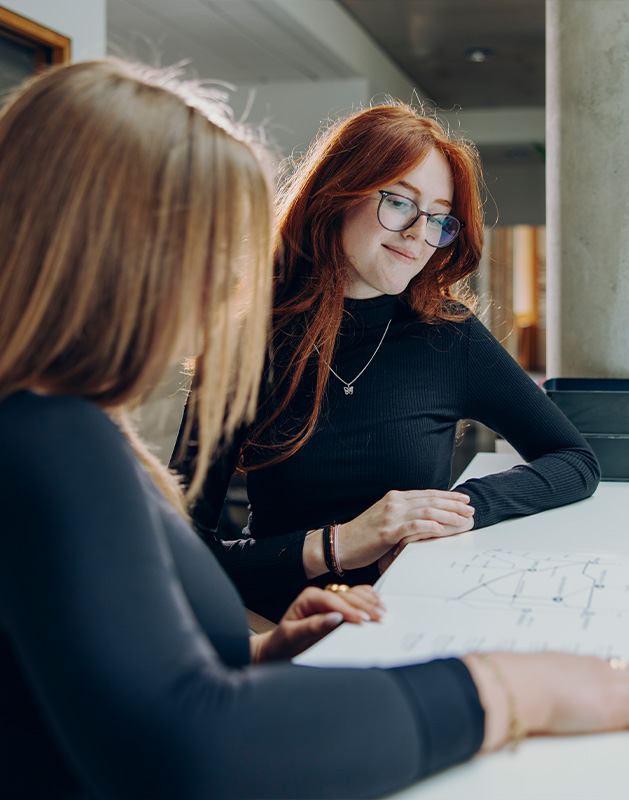 Zwei Frauen mit langen Haaren, eine mit roten Haaren und Brille, betrachten gemeinsam Dokumente am Tisch.