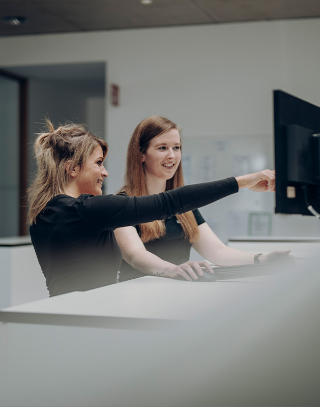 Zwei Frauen, die an einem Computer arbeiten und miteinander sprechen, während sie auf den Bildschirm zeigen.