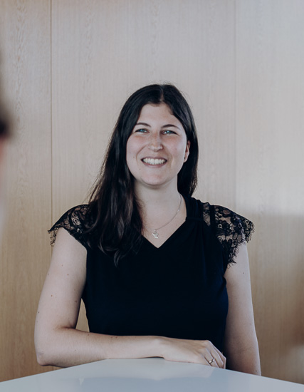 Frau mit langen, dunklen Haaren in einem schwarzen Oberteil mit Spitzenärmeln, lächelnd vor einer Holzoberfläche.