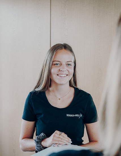 Junge Frau mit langen Haaren in einem schwarzen T-Shirt von SCHORER + WOLF, lächelnd und mit einem Ring an der Hand.