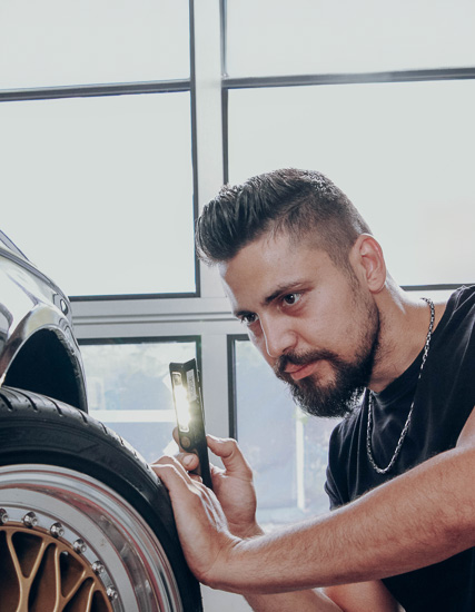 Mann untersucht ein Autoreifen mit einer Taschenlampe in einer Werkstattumgebung.