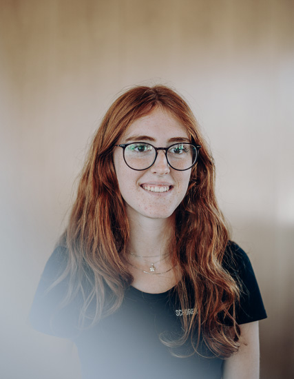 Junge Frau mit langen, roten Haaren und Brille, die in einem hellen Raum steht und lächelt.