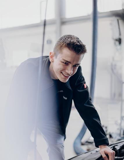 Junger Mann mit kurzem Haar, der an einem Fahrzeug arbeitet und dabei lächelt.