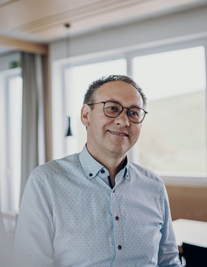 Mann mit Brille und Hemd mit Punkten, lächelnd in einem Innenraum mit Fenstern.