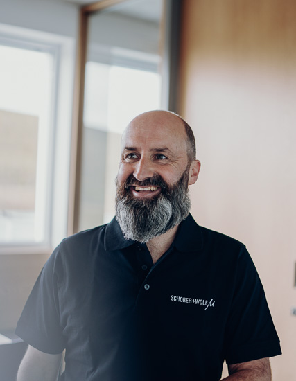 Mann mit Bart in schwarzem Polohemd mit Logo von SCHORER + WOLF, lächelnd in einem hellen Raum.