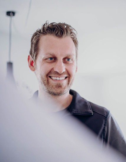Lächelnder Mann mit kurzen, blonden Haaren und Bart, der in einem hellen Raum steht.