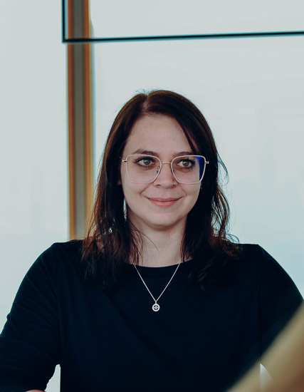 Frau mit Brille und dunklen Haaren, die in einem modernen Raum sitzt und in die Kamera lächelt.