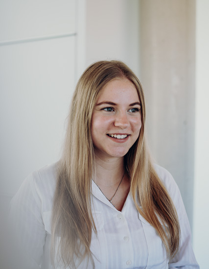 Junge Frau mit langen, blonden Haaren und einem weißen Hemd, lächelnd in einem Innenraum.