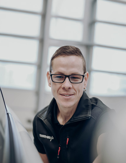 Mann mit Brille und schwarzer Weste, der in einer Werkstatt arbeitet und in die Kamera lächelt.