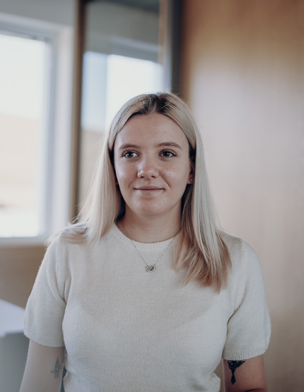 Junge Frau mit blonden Haaren in einem hellen Pullover, steht in einem Raum mit Fenstern.