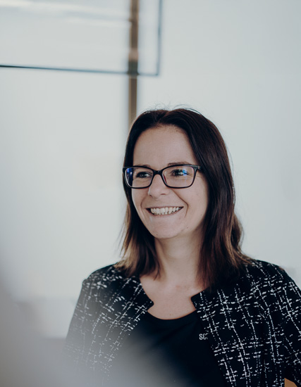 Frau mit Brille und lächelndem Gesicht, trägt eine schwarze Jacke mit Muster, in einem modernen Innenraum.