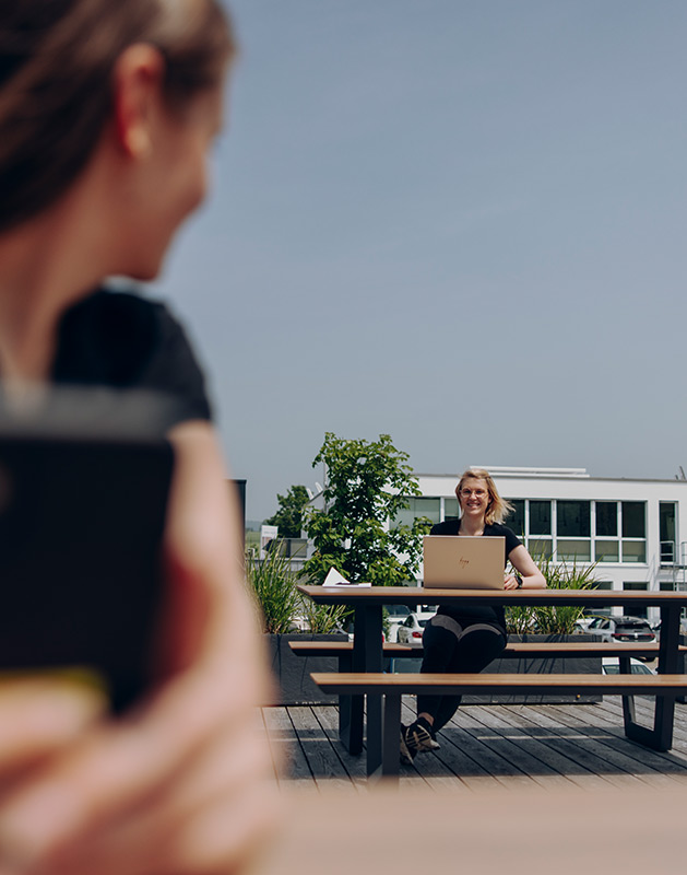 Frau sitzt an einem Tisch im Freien mit einem Laptop und lächelt, während eine andere Person sie anblickt.