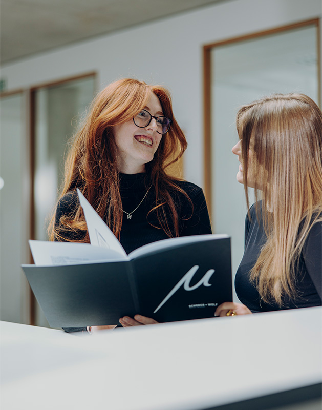 Zwei Frauen mit langen Haaren, die in einem modernen Büro miteinander sprechen und ein Dokument betrachten.