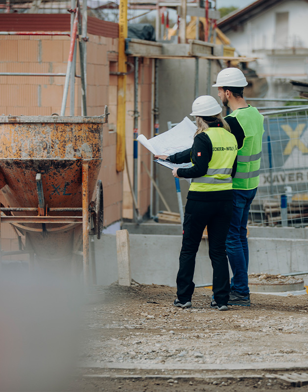 Job-Bauingenieur-_-Sachverständiger-für-Gebäude-und-Objekte Zwei Bauingenieure in Sicherheitswesten und Helmen betrachten Baupläne auf einer Baustelle.