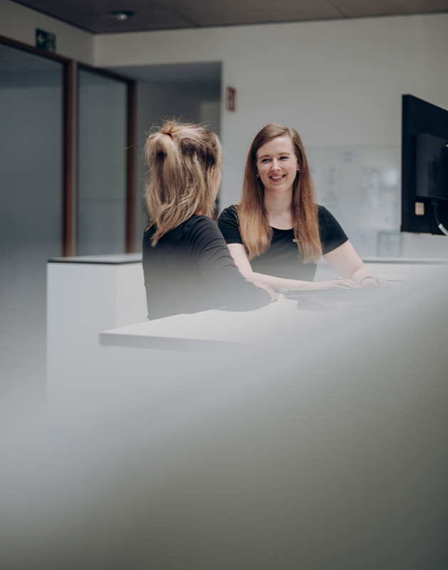 Job-Schnupperpraktikum-Kauffrau-_-Kaufmann-für-Büromanagement Zwei Frauen im Gespräch an einem Empfangstresen in einem modernen Büro mit Computer im Hintergrund.