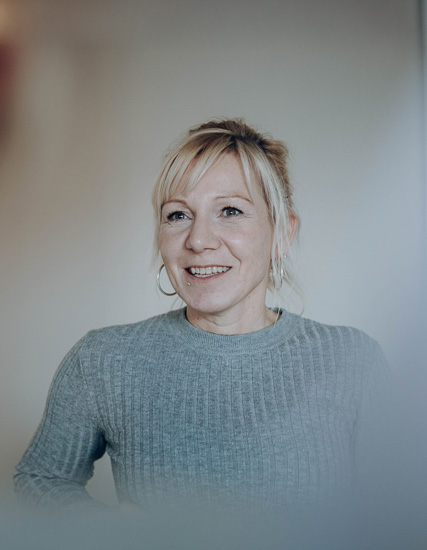 Frau mit blonden Haaren und grauem Pullover, die lächelt und in die Kamera schaut.